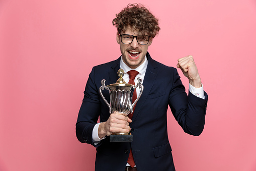 excited young man in navy blue suit wearing glasses and screaming, holding trophy, holding fists up and celebrating victory on pink background