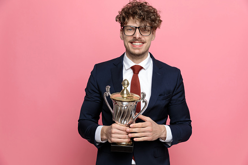 excited young man in navy blue suit wearing glasses, holding trophy and smiling on pink background