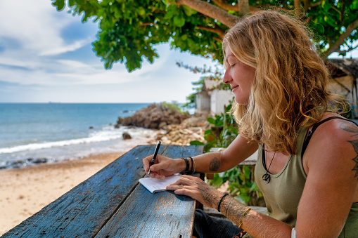 One smiling adult woman is writing on paper with a pen.  She is sitting at a wooden bench overlooking a beach whilst on vacation.
