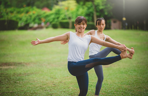 Two Fitness Asian women doing yoga Utthita Hasta Padangusthasana pose (Extended Hand-to-Big-Toe Pose) and relax with sportswear in city public park. Concept of Meditation, Relaxation and Healthy lifestyle.