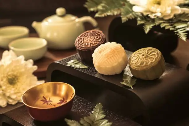 Snowskin Mooncake, the non-traditional mooncake that doesn't require any baking. Sweet bean paste wrapped in mochi dough of various flavours, then moulded in mooncake mould. Three mooncakes of cocoa, milk and matcha flavours are arranged on a wooden serving block. Chrysanthemum flower and fern leaves are used as decoration.