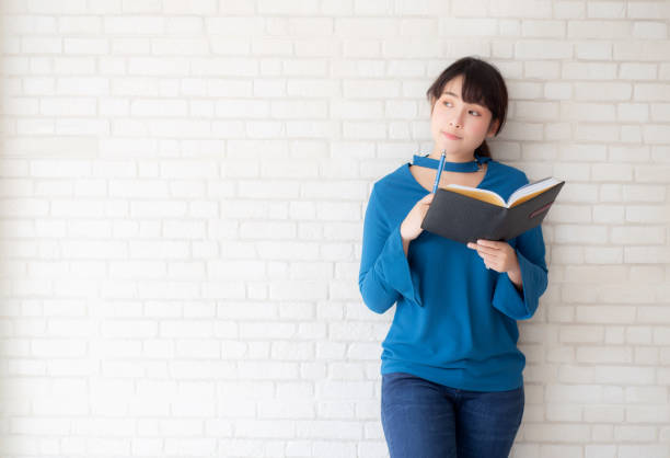 istock Beautiful asian woman smiling standing thinking and writing notebook on concrete cement white background at home, girl homework on book, education and lifestyle concept. 1271146319