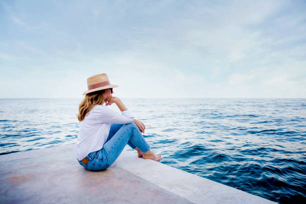 崖の上に座って海を見ながら、リラックスした女性が白いシャツと青いジーンズを着て - mind trip ストックフォトと画像