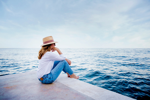 Shot of woman wearing straw hat and sunglasses while sitting on seaside and enjoying the sea view.