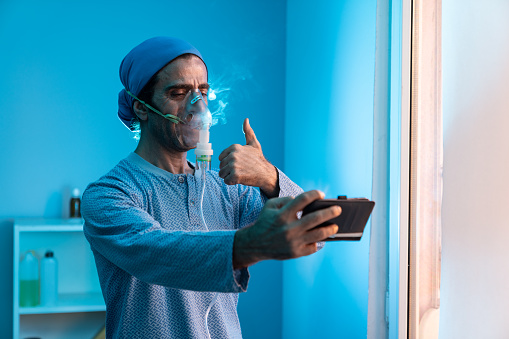 Portrait of mature adult man using nebulizer and using smartphone for video call. He is wearing blue colored hospital clothing. Shot indoor  with a full frame mirrorless camera.