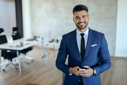Portrait of young happy entrepreneur holding mobile phone and looking at camera while standing in the office.