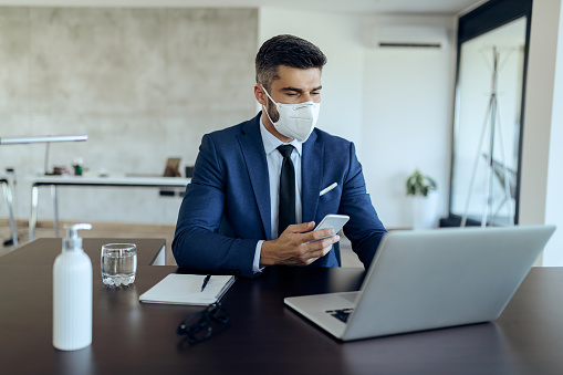 Male entrepreneur working on computer while using mobile phone and wearing face mask due to COVID-19 epidemic.