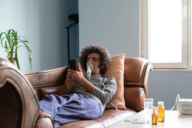 Mature Adult Man Using Nebulizer At Home Mature adult man lying down on sofa in living room and using nebulizer during coronavirus pandemic. He is wearing sleepwear and using smartphone for communication. Shot indoor with a full frame mirrorless camera. medical oxygen equipment stock pictures, royalty-free photos & images