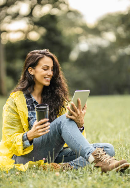 istock Beautiful young lady in yellow raincoat reading a book on her digital tablet, drinking tea and sitting on grass while taking a break in nature 1271165714