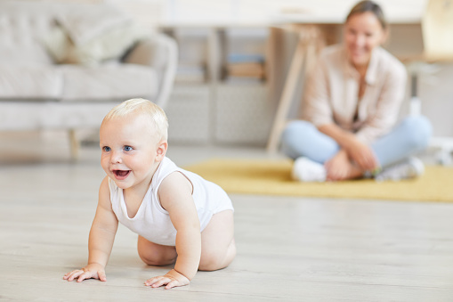 Young woman watching her cheerful little son crawling on his fours at home, copy space