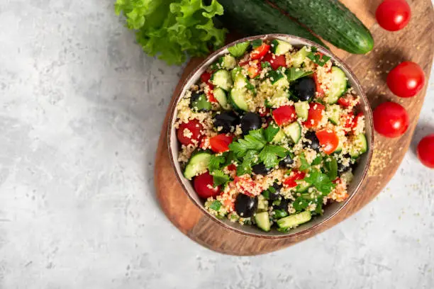Salad with couscous and vegetables in a bowl on a gray concrete background top view. Copy space. Tabbouleh of couscous, tomatoes, cucumbers, parsley and olives. Arabic or African cuisine.