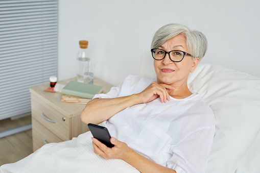 Portrait of beautiful aged woman wearing white clothes lying on bed in hospital ward holding smartphone looking at camera