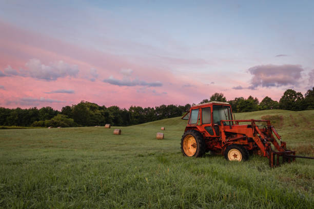 Red tractor and freshly rolled hay bales rest on rolling hill with dramatic cloudscape at sunrise.  Rustic rural scene with copyspace.