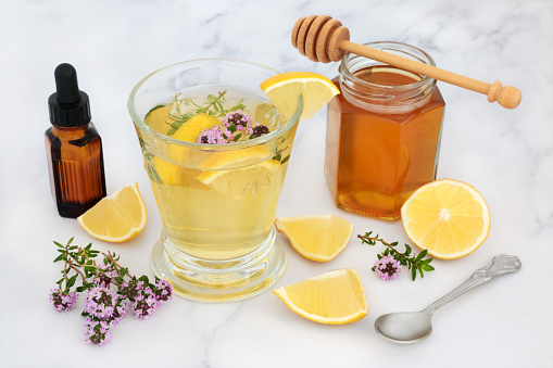 Alternative medicinal remedy for cold & flu virus with thyme herb, honey & lemon in glass with essential oil. Immune boosting medication, is anti bacterial, antiseptic and anti viral. On marble background.