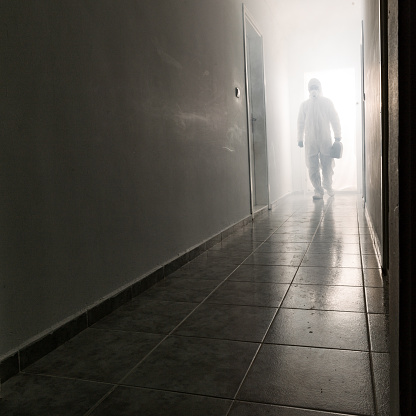 Man wearing protective hazmat suit, protective gloves, face mask and eyewear walking in corridor. The suit is white. The corridor is foggy. He is seen in full length and holding a hard case. Shot indoor with a medium format camera during covid-19 pandemic.