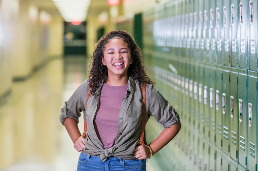 An African-American high school student standing in the hallway of her school by a row of lockers. She is a teenage girl, 16 years old, carrying a backpack, looking at the camera, smiling.