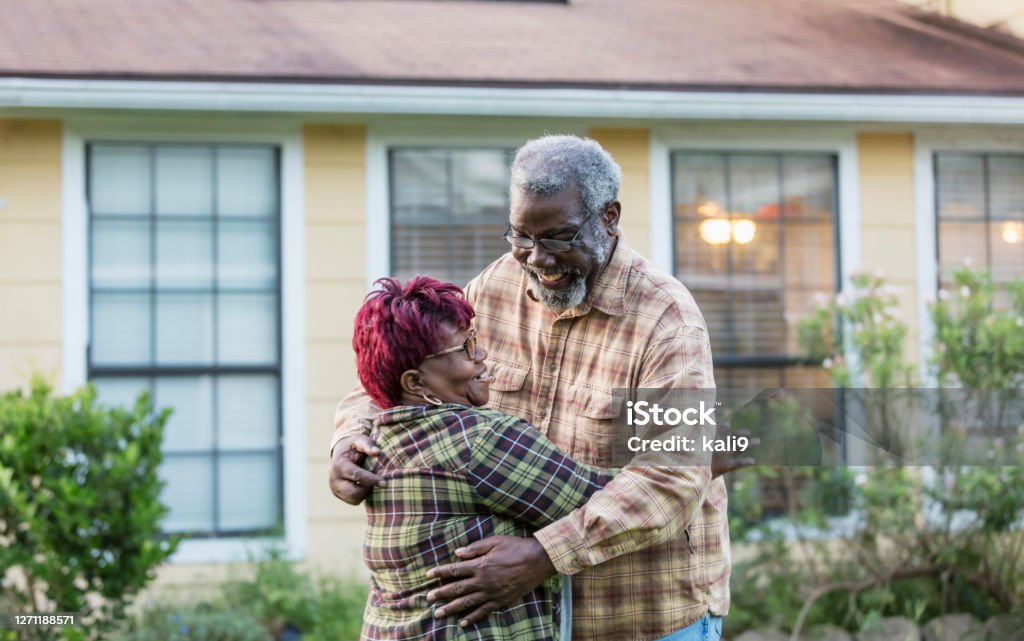 Senior African-American couple hugging in front of home A senior African-American couple, in their 70s, standing together outside their home, in the front yard, smiling as they embrace each other. House Stock Photo