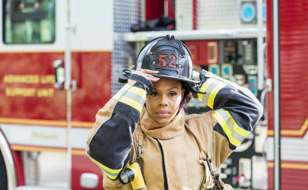 Female firefighter in fire protection suit An African-American female firefighter wearing a fire protection suit, standing next to a fire engine. She is a mid adult woman in her 30s with a serious expression. firefighter stock pictures, royalty-free photos & images