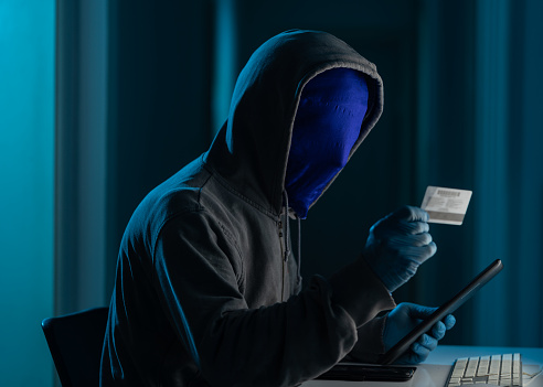 Photo of adult man wearing gloves, a gray hooded sweater to hide his identity and using computer for hacking.The background is dark and atmosphere is futuristic. He is holding a credit card and a digital tablet while typing.