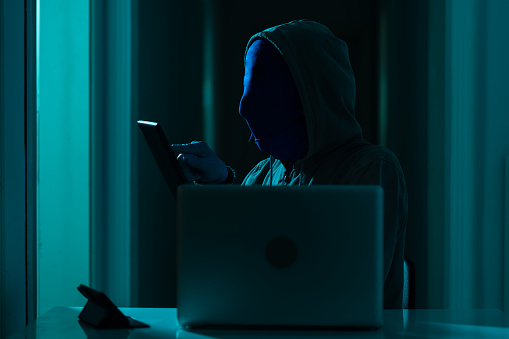 Photo of adult man wearing gloves, a gray hooded sweater to hide his identity and using computer for hacking.The background is dark and atmosphere is futuristic. He is holding a a smartphone while typing.