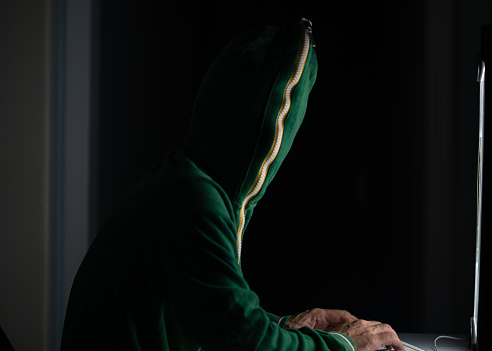Photo of adult man wearing a green zipped hooded sweater to hide his identity and using computer for hacking.The background is dark and atmosphere is futuristic. He is using a keyboard