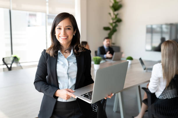 Smiling female entrepreneur with laptop in a meeting room Portrait of smiling female entrepreneur holding a laptop with team in background at office conference room businesswoman stock pictures, royalty-free photos & images