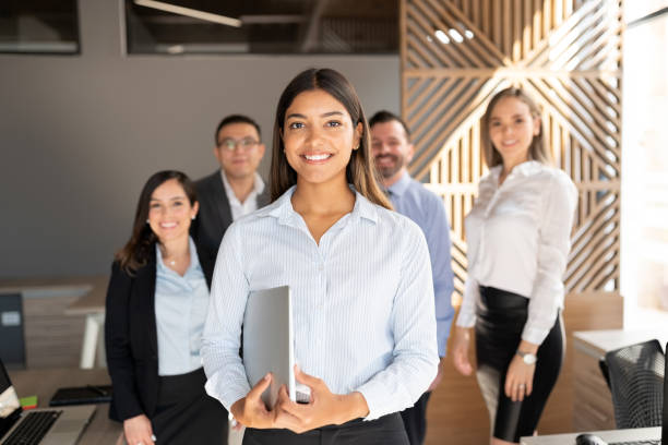 Confident hispanic business woman in office Confident hispanic businesswoman standing in office with colleagues in background latin american and hispanic ethnicity stock pictures, royalty-free photos & images