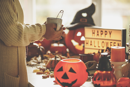 Cut out of female hands holding cup of tea with seasonal decorative items and Happy Halloween sign in the backgroung