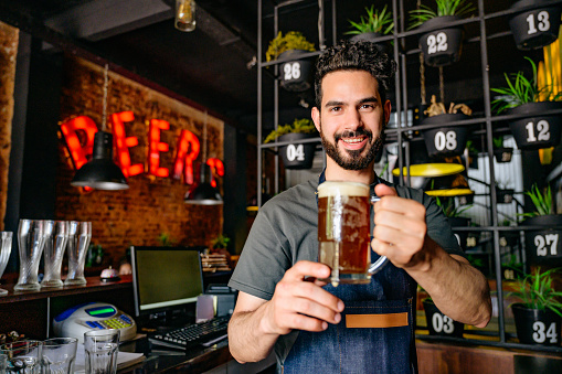 Close-up of bearded bartender in mid 20s holding up mug of beer fresh from the tap at Buenos Aires bar.