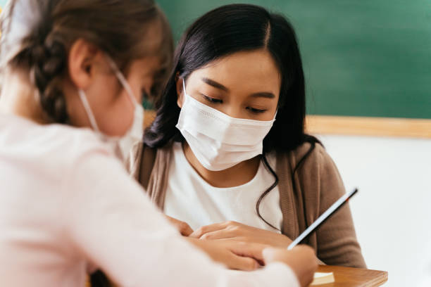 Close-up of Asian female teacher wearing a face mask in school building tutoring a primary student girl. Elementary pupil is writing and learning in classroom. Close-up of Asian female teacher wearing a face mask in school building tutoring a primary student girl. Elementary pupil is writing and learning in classroom. Covid-19 school reopen concept tutor stock pictures, royalty-free photos & images