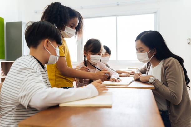 学校の舎の多様な生徒のグループがグループ学習クラスで議論します。小学生はブレーンストーミング中にフェイスマスクを着用しています。 - elementary school building ストックフォトと画像