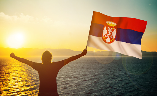 The man is waving the Serbian Flag against the sunrise. Patriotism concept. Horizontal composition with copy space.