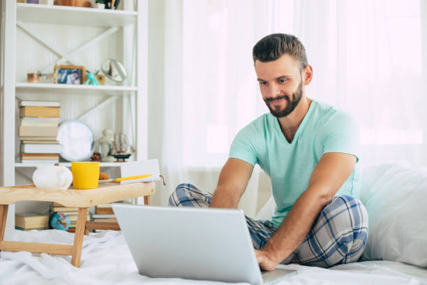 handsome young bearded man in casual clothes is working at home while sitting on the bed. confident guy with a laptop and smartphone drinks coffee in bedroom. - business styles foods and drinks drinking imagens e fotografias de stock