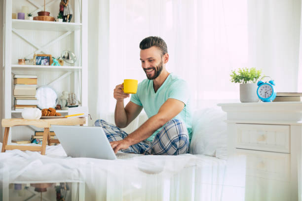 istock Handsome young bearded man in casual clothes is working at home while sitting on the bed. Confident guy with a laptop and smartphone drinks coffee in bedroom. 1271264211