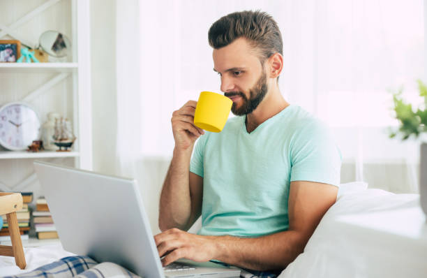 istock Handsome young bearded man in casual clothes is working at home while sitting on the bed. Confident guy with a laptop and smartphone drinks coffee in bedroom. 1271264225
