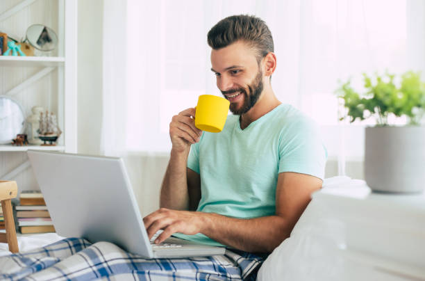 istock Handsome young bearded man in casual clothes is working at home while sitting on the bed. Confident guy with a laptop and smartphone drinks coffee in bedroom. 1271264232