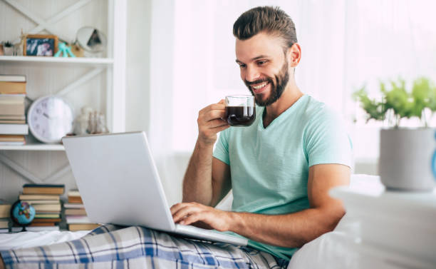 istock Handsome young bearded man in casual clothes is working at home while sitting on the bed. Confident guy with a laptop and smartphone drinks coffee in bedroom. 1271264253