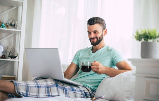 istock Handsome young bearded man in casual clothes is working at home while sitting on the bed. Confident guy with a laptop and smartphone drinks coffee in bedroom. 1271265162