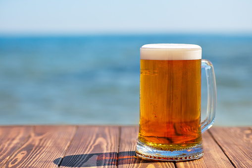 mug of light beer with froth and bubbles on wooden table with sea on the background