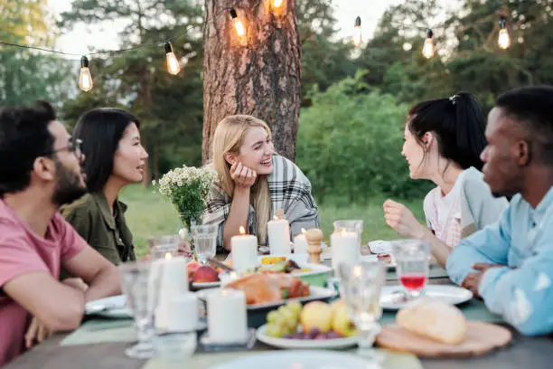 Photo of Young joyful couples eating and talking by served festive table under pine tree