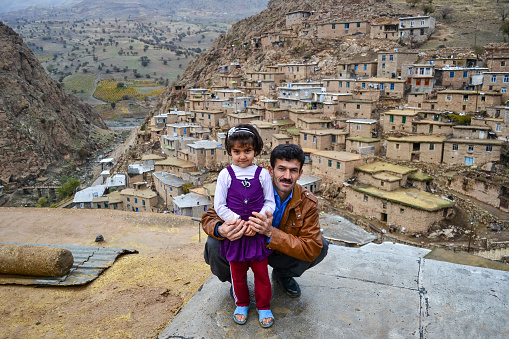 Palangan, Iranian Kurdistan - November 15, 2013: Portrait of cute and little Kurdish girl huged by her father while posing with Palangan stone village in the background
