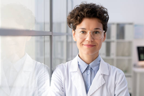 Young successful female worker of scientific lab in whitecoat and eyeglasses Young successful female worker of scientific laboratory in whitecoat and eyeglasses standing by glass wall inside office in front of camera scientist stock pictures, royalty-free photos & images