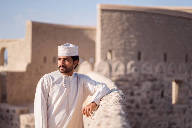 bonito jovem muçulmano omani com roupas tradicionais thwab brancas e chapéu muçulmano posando no forte bahla - bahla fort - fotografias e filmes do acervo