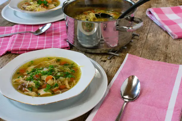 fresh and homemade chicken soup with noodles and vegetables served on a white plate on a wooden table with soup pot and ladle - ready to eat