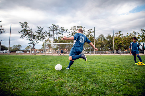 Rear view of 12 year old Caucasian male athlete wearing blue uniform and approaching ball for kick toward distant goal.