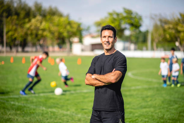 Portrait of Male Footballer Coaching Mixed Age Players Mature male coach standing with arms crossed and looking at camera on sports field as young athletes practice in background. 6 11 months stock pictures, royalty-free photos & images