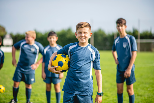 Smiling pre-teen boy athlete holding soccer ball and smiling at camera as teammates stand in background.
