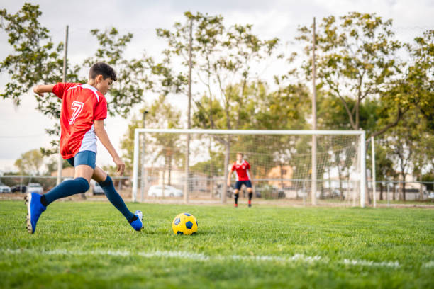 Athletic Boy Footballer Practicing Kick to Goal Low angle view of 14 year old male athlete taking practice kicks with goalie in background ready to defend. teen goalie stock pictures, royalty-free photos & images