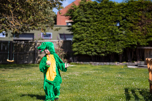 A medium shot of a young, Caucasian boy running with a water balloon during a water fight in his back garden in Northeastern England. The boy is also dressed in a dinosaur costume.