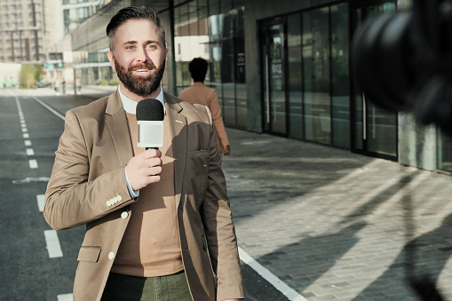 Mature bearded man speaking in microphone and smiling at camera during live broadcasting on city street